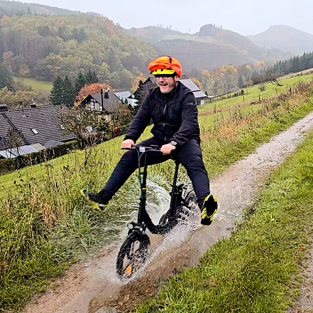 An action-packed scene shows a rider splashing through a muddy countryside trail on the iScooter U4 electric bike, surrounded by rolling hills and scenic mountain views. The image highlights the low step-through frame, sturdy wheels, front suspension, and upright riding position, showcasing the iScooter U4's versatility and comfortable performance on both urban roads and outdoor paths.
