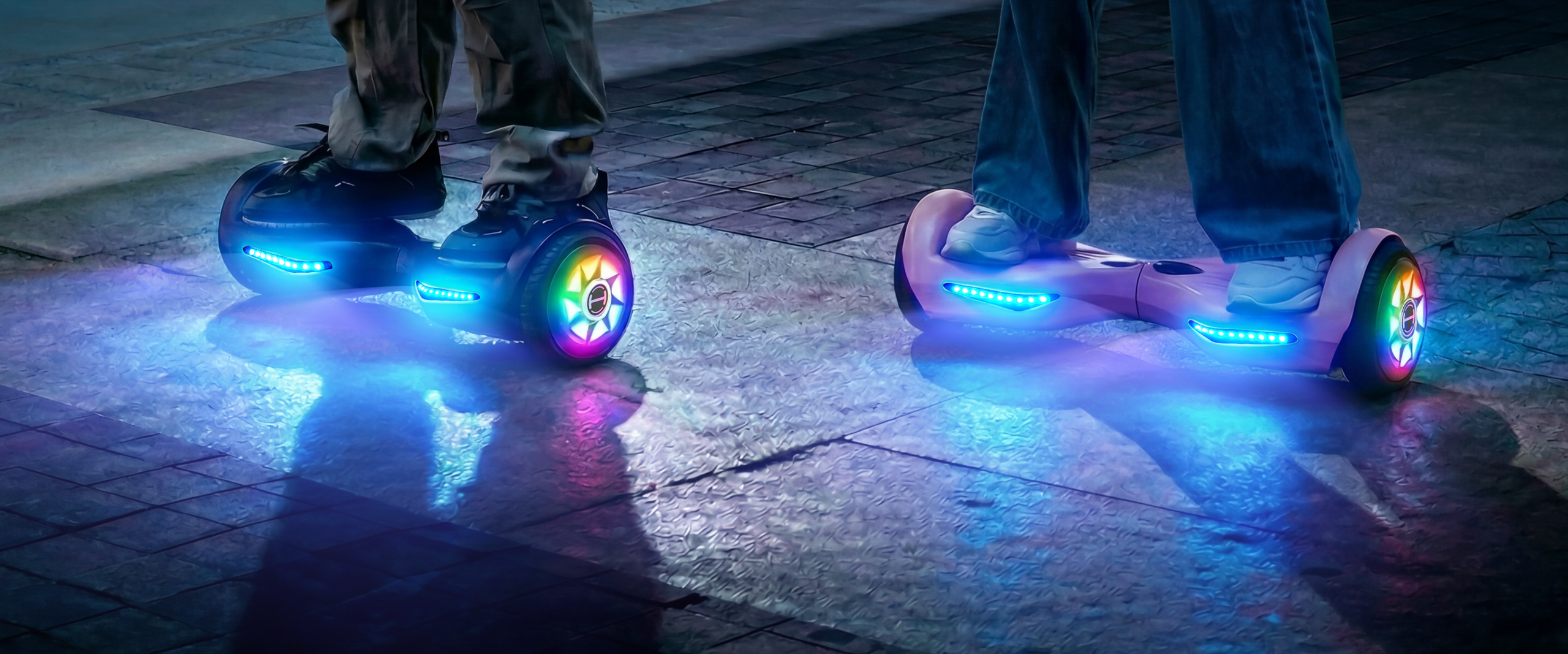 Two riders stand on iHoverboard H1 hoverboards at night, each with bright LED lights illuminating the wheels and foot platforms, casting colorful reflections on the ground. One hoverboard is a sleek dark design, while the other is a light pink model. The scene highlights the H1’s futuristic look, glowing style, and playful nighttime riding experience.