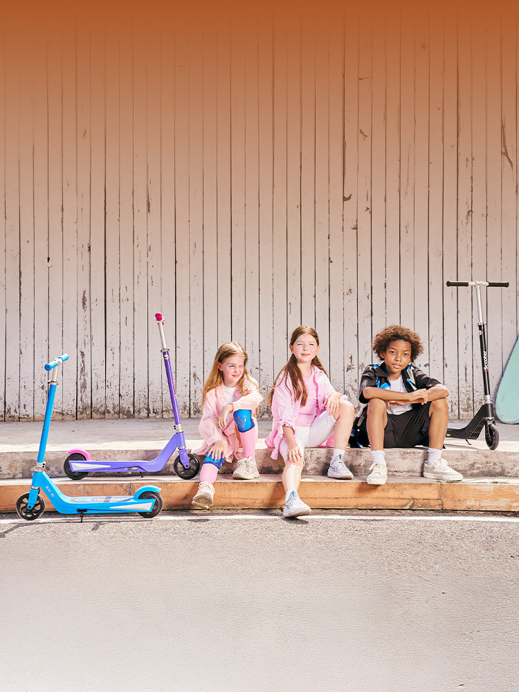 Three young friends relaxing outdoors next to colorful iScooter IK1S electric scooters designed for kids. Crafted for young riders, this beginner-friendly model incorporates smooth-acceleration controls, a durable non-slip deck, vibrant color choices, and a lightweight frame that makes daily neighborhood riding safe and effortlessly portable.