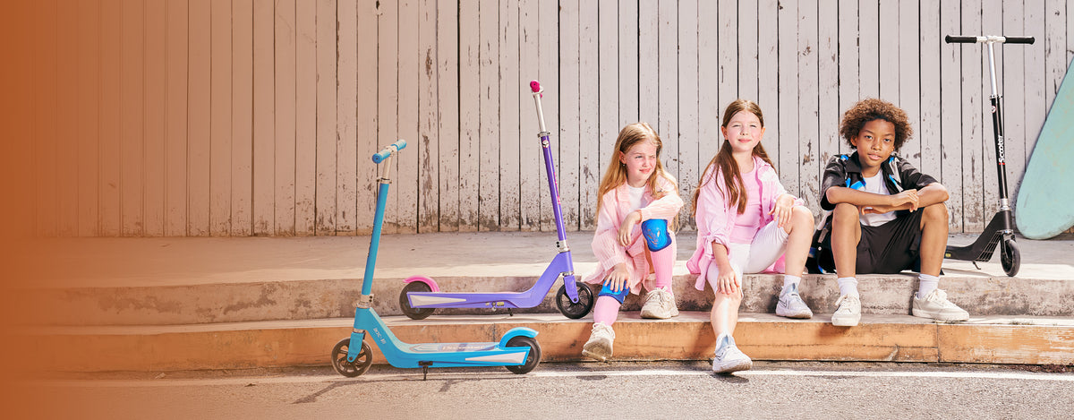 Group of young children sitting on a ledge alongside vibrant iScooter IK1S kids electric scooters displayed in multiple bright colors. Designed specifically for beginners, this lightweight youth e-scooter features child-friendly handlebars, an easy-to-use foot pedal accelerator, safe low-speed limits, and a sturdy deck to give kids a fun, safe entry into electric riding.