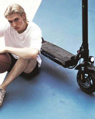 Lifestyle shot of a young man holding a basketball while sitting on a blue sports court next to an iScooter iX3 electric scooter. Highlighting front disc brakes, front hydraulic shock absorption, a bright LED headlight, and a wide textured standing deck, this stylish e-scooter brings sporty versatility and reliable comfort to urban athletic lifestyle activities.