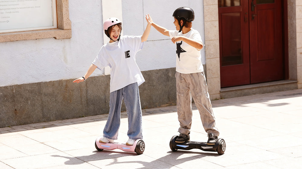Heartwarming lifestyle scene of two young kids high-fiving while riding self-balancing hoverboards on a sunny sidewalk, with the girl on a pastel pink iHoverboard H1. Tailored for children and beginner riders, this easy-to-use hoverboard features colorful LED wheel lights, anti-slip footpads, front safety headlights, and intelligent self-balancing technology for a safe, fun, and confident riding experience.