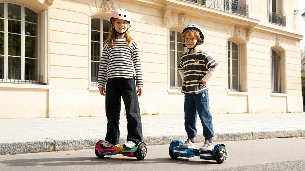 Charming outdoor lifestyle scene depicting two young children wearing protective helmets standing on self-balancing hoverboards outside classic European architecture, with the boy riding a metallic blue iHoverboard H2. Perfect for kids and young beginners, this fun hoverboard features LED-lit wheel hubs, non-slip rubber footpads, integrated safety sensors, and smooth self-balancing control for stable recreational riding.