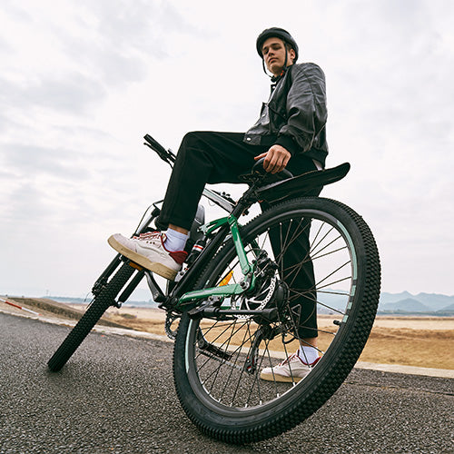 Rider seated on rugged iHoverboard M50 electric mountain bike with integrated battery pack, responsive suspension, and disc brakes, captured from low angle on paved road with scenic hills in background. The M50 delivers extended range, powerful acceleration, and smooth off‑road handling — making it ideal for adventurous outdoor cycling.
