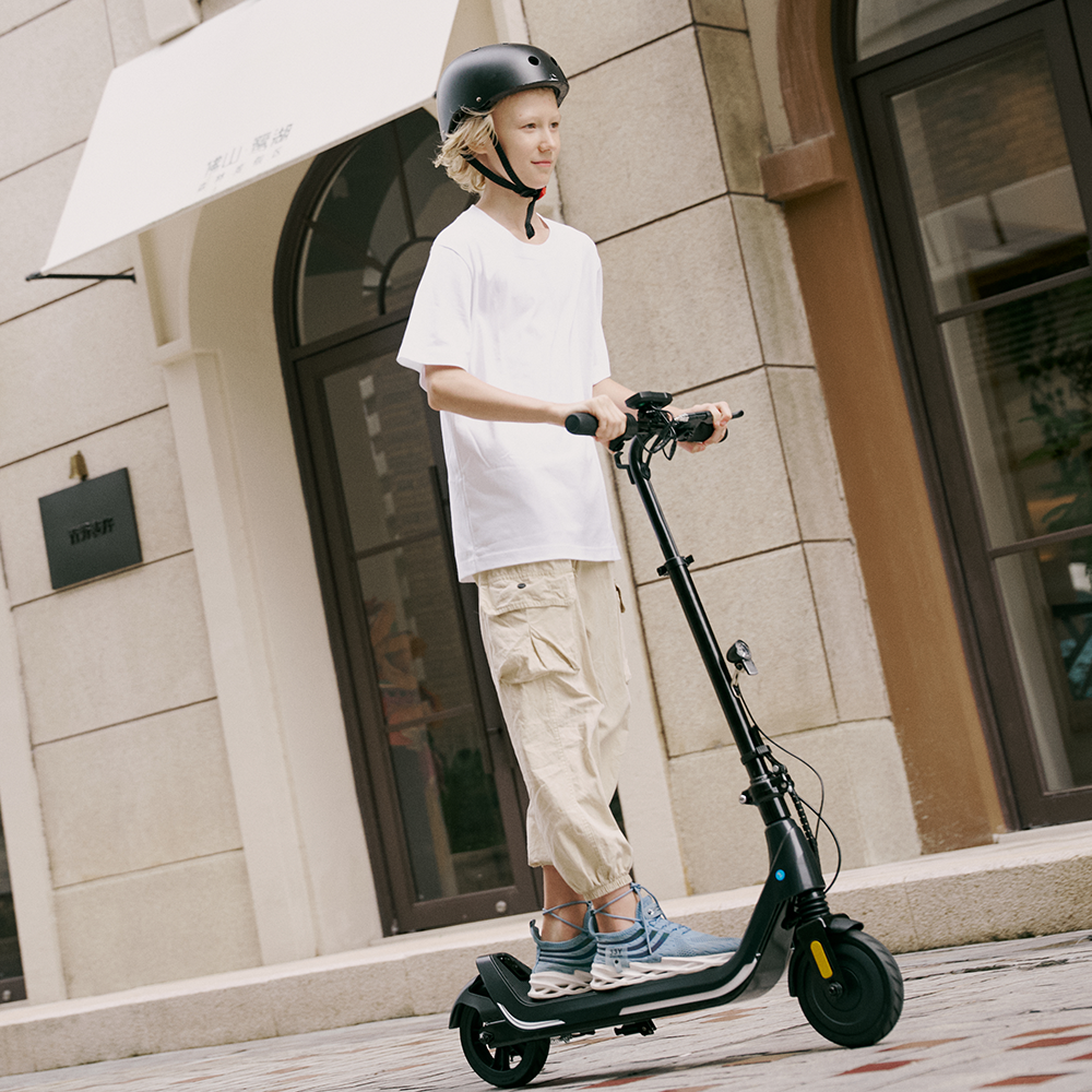 Boy wearing a helmet riding the black iScooter iK5 electric scooter on a paved walkway, emphasizing its responsive hand controls, comfortable foot platform, front suspension system, and reliable traction for outdoor cruising.