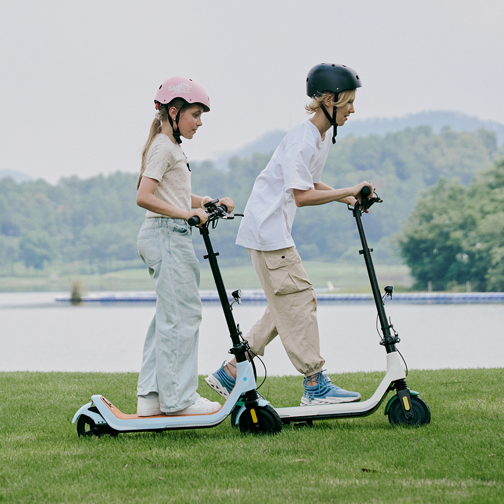 Two young adventurers wearing helmets gliding side by side on light blue and white iScooter iK5 electric scooters along a scenic park path near a lake, highlighting the stable wide foot decks, reliable dual-wheel stability, adjustable height handlebars, and smooth ride quality tailored for outdoor family fun.
