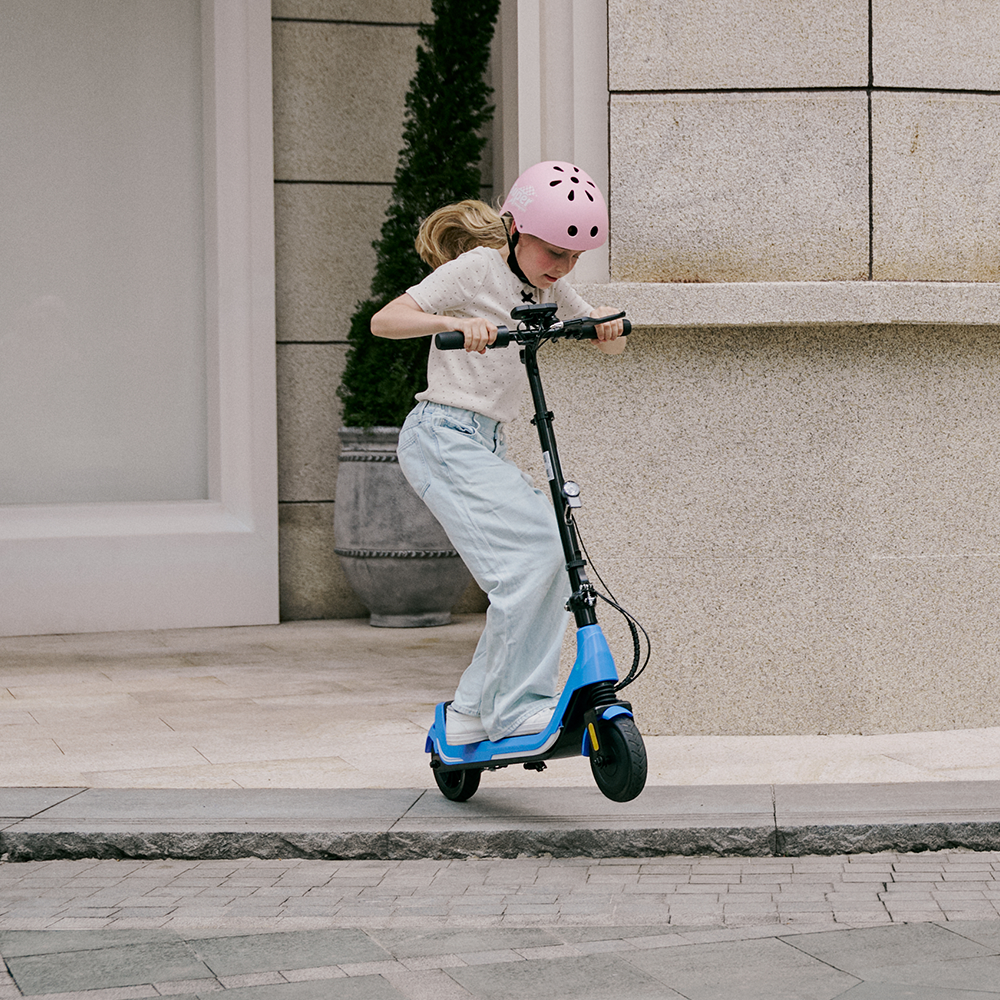 Young rider wearing a pink safety helmet performing a front wheel lift on the blue iScooter iK5 electric scooter, showcasing its durable construction, responsive design, powerful motor, and ergonomic handlebars perfect for kids practicing tricks and active play on paved urban streets.