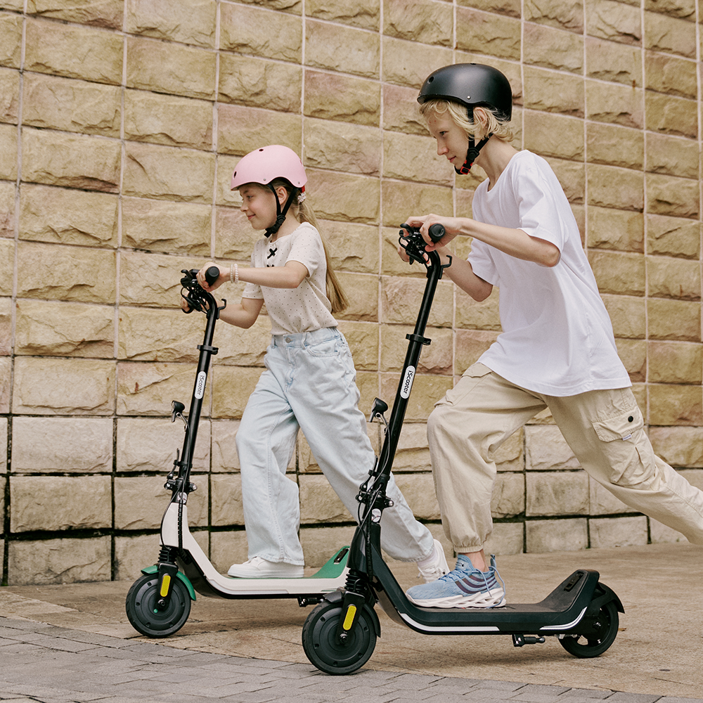 Two kids wearing helmets cruising alongside a stone wall on white and black iScooter iK5 electric scooters, displaying their sturdy build, comfortable wide decks, safety reflectors, and dual front suspension designed for fun and safe outdoor rides.