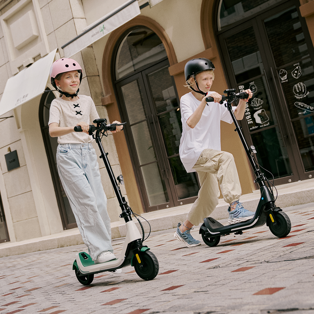 Pair of young riders equipped with helmets navigating a paved town square on white and black iScooter iK5 electric scooters, emphasizing their sturdy frame structures, front wheel shock absorption, easy-grip handlebars, and reliable braking performance tailored for active play.