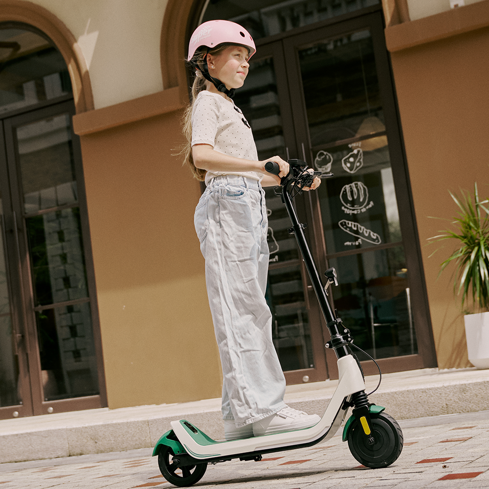 Confident young girl wearing a pink safety helmet riding the white and green iScooter iK5 electric scooter outside a storefront, highlighting its sleek body frame, reliable integrated braking, stable standing deck, and smooth wheels designed for effortless neighborhood travel.