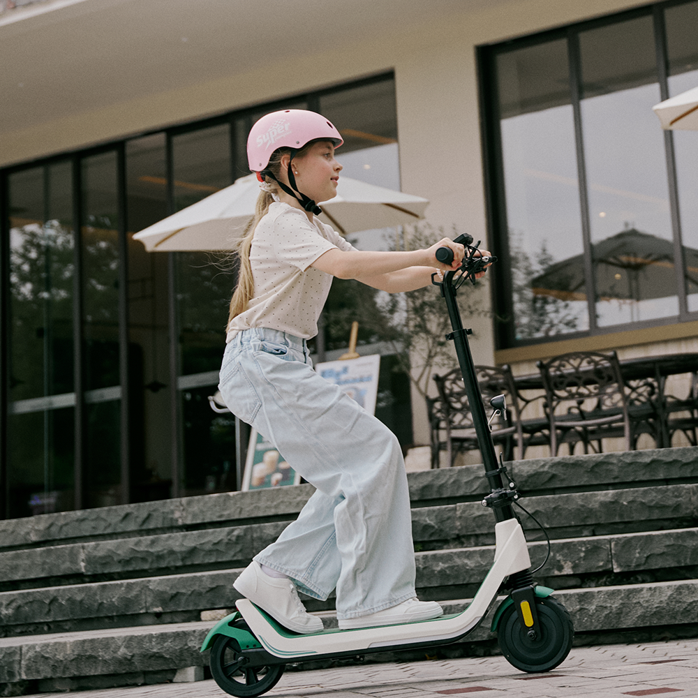 Enthusiastic young girl in a pink helmet riding a white and green iScooter iK5 electric scooter across a paved walkway, emphasizing its low-profile deck, comfortable standing space, front shock absorption, and secure handlebar grips for daily riding adventures.