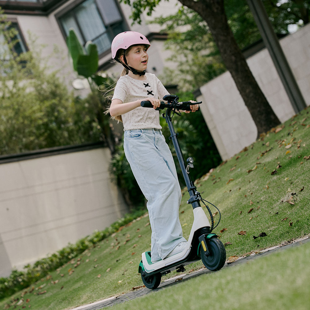 Active young girl wearing a pink safety helmet steering a white and green iScooter iK5 electric scooter along an outdoor stone path, featuring its upright handlebar assembly, integrated LED headlight, sturdy baseboard, and puncture-resistant tires built for reliable neighborhood exploration.