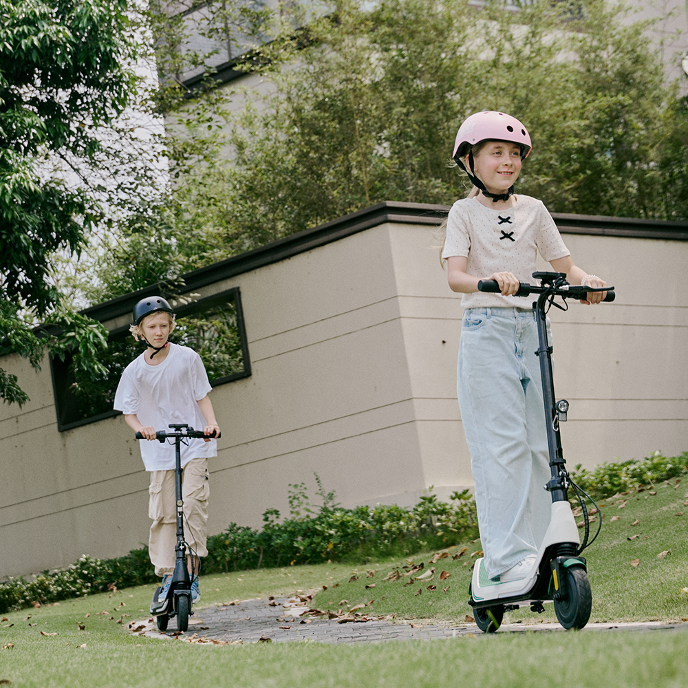Smiling young girl wearing a protective pink helmet leading a friend on a stone walkway riding the white and green iScooter iK5 electric scooter, showcasing its front LED headlight, sturdy frame, wide footing platform, and smooth treaded wheels perfect for outdoor recreational adventures.